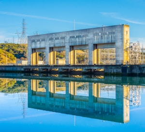 A large concrete hydroelectric power station structure is reflected in a calm body of water. The building features multiple large, square openings and is surrounded by electrical infrastructure, including visible power lines and towers. The sky is clear with a bright blue tone, suggesting a sunny day. Hills and greenery are visible in the background.