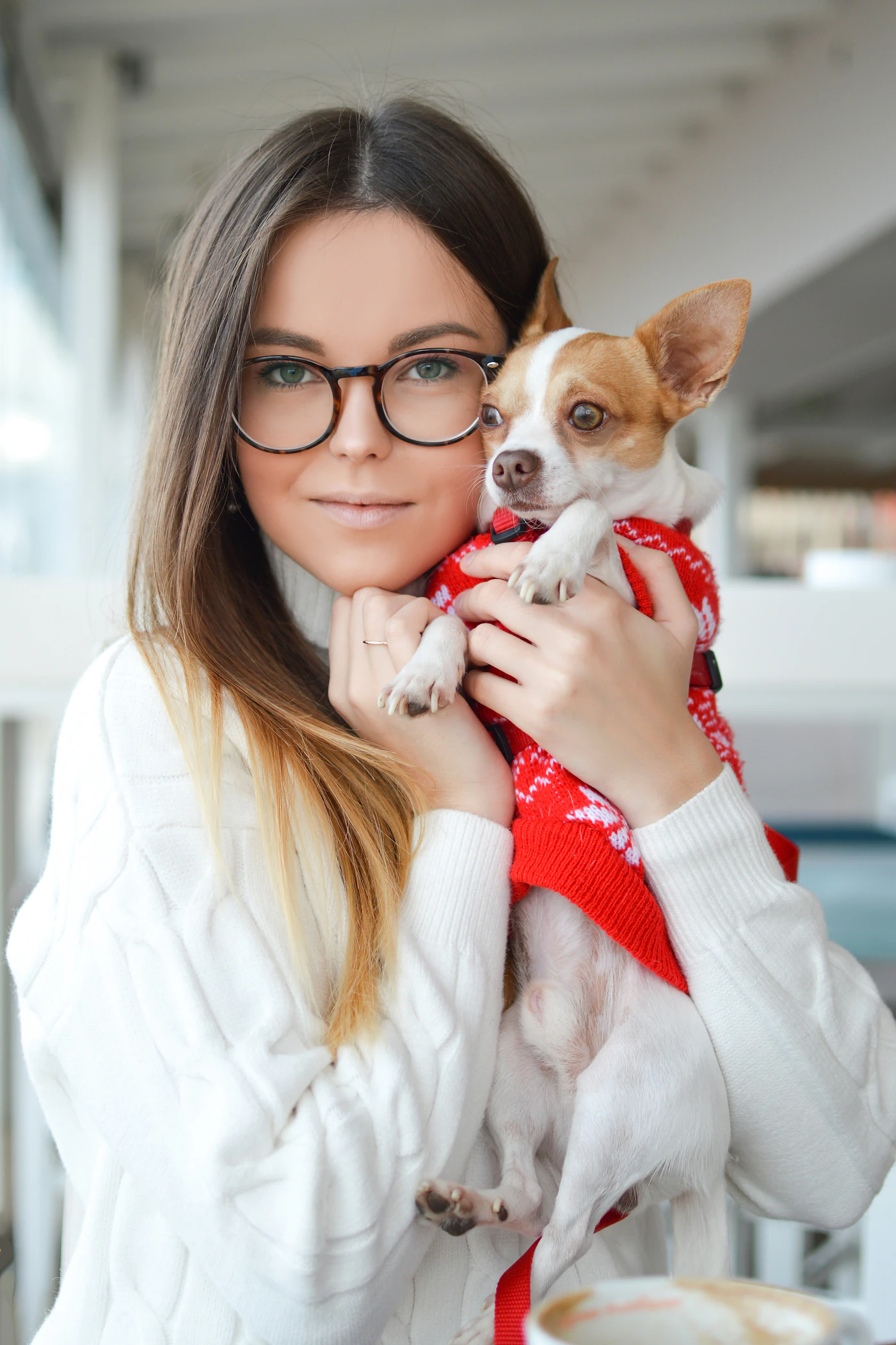 A woman in glasses and a white sweater gently holding a small dog wearing a red knit sweater