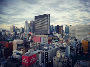 An urban landscape featuring a densely populated cityscape with high-rise buildings and a skyline. The architecture includes modern skyscrapers and smaller structures scattered below. A variety of colors from billboards and advertisements add vibrancy to the scene. Clouds fill the sky, adding texture to the backdrop.
