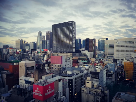 An urban landscape featuring a densely populated cityscape with high-rise buildings and a skyline. The architecture includes modern skyscrapers and smaller structures scattered below. A variety of colors from billboards and advertisements add vibrancy to the scene. Clouds fill the sky, adding texture to the backdrop.
