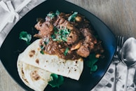 A plate featuring a brown meat curry garnished with fresh green cilantro, accompanied by two pieces of flatbread. The setting includes a folded fabric napkin with cutlery placed beside the plate, all arranged on a wooden table.