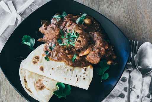 A plate featuring a brown meat curry garnished with fresh green cilantro, accompanied by two pieces of flatbread. The setting includes a folded fabric napkin with cutlery placed beside the plate, all arranged on a wooden table.