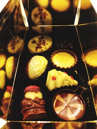 A box containing an assortment of chocolates, including shapes like a Santa figure, a Christmas tree, and various filled and decorated pieces. These chocolates have been carefully arranged with intricate designs including some with red accents.