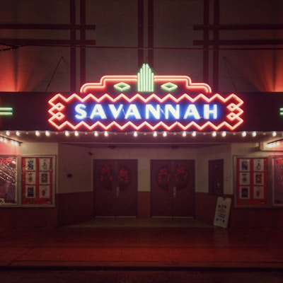 A vintage theater facade brightly lit with neon lights. The marquee displays the name 'Savannah' in bold red and blue neon lettering. The building features art deco elements with geometric patterns surrounding the sign. Posters and advertisements can be seen in the display cases on either side of the entrance doors, and a sandwich board sign is positioned on the sidewalk.