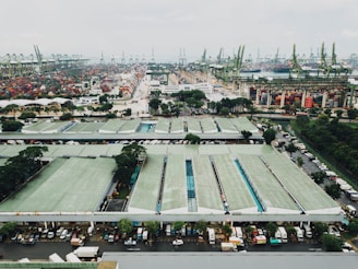 A large industrial port area with numerous shipping containers neatly arranged in rows. Several cranes are positioned around the area, likely for loading and unloading cargo from ships. In the foreground, there are warehouse buildings with green roofs and a line of vehicles, including trucks, parked along the side. The background features a view of the sea with more cranes and containers visible, stretching out into the distance.