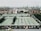A large industrial port area with numerous shipping containers neatly arranged in rows. Several cranes are positioned around the area, likely for loading and unloading cargo from ships. In the foreground, there are warehouse buildings with green roofs and a line of vehicles, including trucks, parked along the side. The background features a view of the sea with more cranes and containers visible, stretching out into the distance.