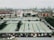 A large industrial port area with numerous shipping containers neatly arranged in rows. Several cranes are positioned around the area, likely for loading and unloading cargo from ships. In the foreground, there are warehouse buildings with green roofs and a line of vehicles, including trucks, parked along the side. The background features a view of the sea with more cranes and containers visible, stretching out into the distance.