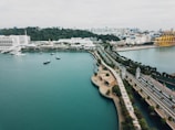 Aerial view of a coastal area with a prominent roadway, lush greenery, and a few boats on the water. The foreground features a wide expanse of water, while the background has urban infrastructure including buildings and industrial structures. The landscape includes a blend of natural and human-made elements.