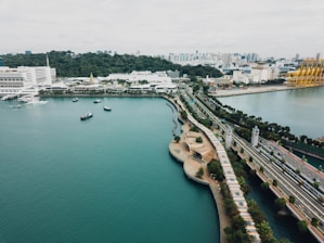 Aerial view of a coastal area with a prominent roadway, lush greenery, and a few boats on the water. The foreground features a wide expanse of water, while the background has urban infrastructure including buildings and industrial structures. The landscape includes a blend of natural and human-made elements.