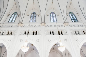 The interior of a church with pristine white walls and large stained glass windows. Elegant archways and ornate light fixtures hang from the ceiling, adding to the grandeur of the space.