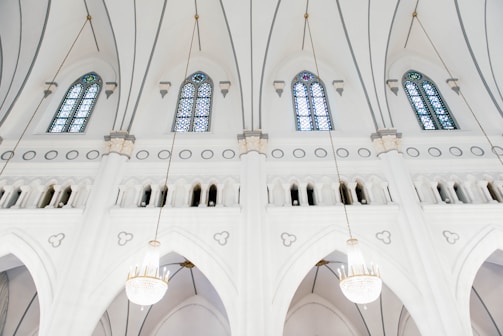 The interior of a church with pristine white walls and large stained glass windows. Elegant archways and ornate light fixtures hang from the ceiling, adding to the grandeur of the space.