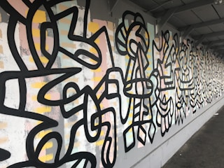 A wall covered in vibrant street art featuring abstract geometric patterns and bold black lines. The background consists of pastel colors such as pink, yellow, and light blue mixed with white and gray tones. The art extends along the wall, creating a continuous and dynamic visual experience.