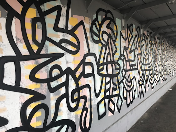 A wall covered in vibrant street art featuring abstract geometric patterns and bold black lines. The background consists of pastel colors such as pink, yellow, and light blue mixed with white and gray tones. The art extends along the wall, creating a continuous and dynamic visual experience.