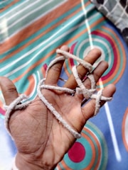 A hand is shown with several light-colored strings loosely wrapped around the fingers. The background consists of a brightly patterned fabric with circular and linear designs in shades of teal, orange, and white.