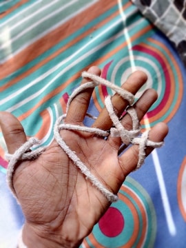 A hand is shown with several light-colored strings loosely wrapped around the fingers. The background consists of a brightly patterned fabric with circular and linear designs in shades of teal, orange, and white.