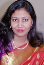 A woman with long, dark hair looks directly at the camera. She is wearing a colorful saree with golden patterns, adorned with a pearl necklace and matching earrings. Her expression is calm and poised.