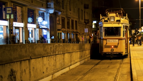 A vintage tram travels along a night-time city street lined with brightly lit shops and buildings. The tram tracks run parallel to a concrete barrier, and there are a few people walking on the side.