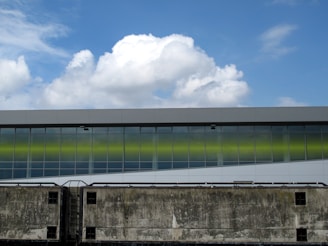 An industrial building facade with a modern design is characterized by a series of large windows reflecting a cloudy sky. The lower half of the image shows a worn, textured wall with pipes running across it, contrasting with the smooth surface of the structure above. A blue sky with fluffy white clouds fills the upper part of the scene.
