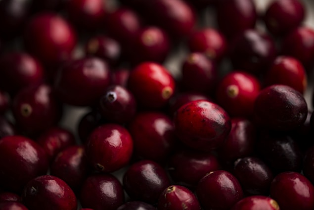 A close-up view of several fresh, smooth cranberries, with a glossy texture and deep red color. The berries are tightly packed together, filling the frame and emphasizing their round, plump shapes.