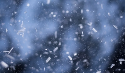 A flurry of snowflakes falls against a blurred backdrop, creating a dreamy, wintry scene. The snowflakes are in various stages of descent, and the soft focus emphasizes the movement and stillness simultaneously.
