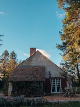 A quaint cottage with a steep, shingled roof and a tall chimney, surrounded by lush green trees. The building features red wooden shutters and a white door with glass panes. In front of the house, a simple wooden table with chairs sits on a brick patio, with lavender plants in the foreground.