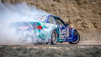 A blue and green sports car drifting on a dirt track, surrounded by a cloud of smoke from the tires. The background features a rocky surface, suggesting an outdoor setting. The car has various decals and numbers, indicating it's participating in a racing event.