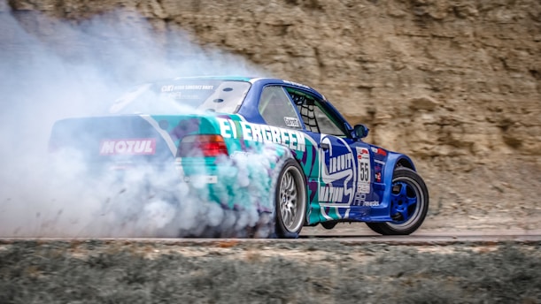 A blue and green sports car drifting on a dirt track, surrounded by a cloud of smoke from the tires. The background features a rocky surface, suggesting an outdoor setting. The car has various decals and numbers, indicating it's participating in a racing event.