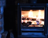 A cozy scene with a wood-burning stove, showcasing actively burning logs and bright flames. The stove is surrounded by brick on one side, with a pile of firewood visible next to it.