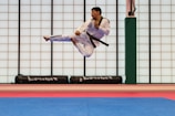 A martial artist performing a high kick in mid-air inside a dojo, wearing a white gi with a black belt. The background is a grid of translucent panels and training mats cover the floor.