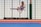 A martial artist performing a high kick in mid-air inside a dojo, wearing a white gi with a black belt. The background is a grid of translucent panels and training mats cover the floor.