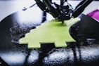 A 3D printer is in the process of creating a lime green object with a textured surface. The object being printed has a distinctive shape, possibly resembling a simplified dinosaur or creative design. The printer appears to be working on a circular black platform, reflecting light due to its glossy surface.