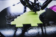 A 3D printer is in the process of creating a lime green object with a textured surface. The object being printed has a distinctive shape, possibly resembling a simplified dinosaur or creative design. The printer appears to be working on a circular black platform, reflecting light due to its glossy surface.