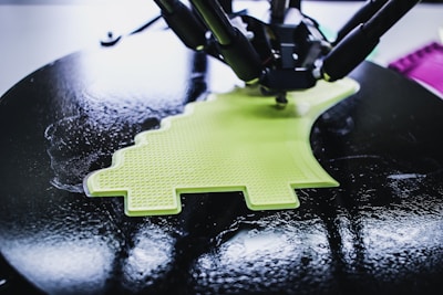 A 3D printer is in the process of creating a lime green object with a textured surface. The object being printed has a distinctive shape, possibly resembling a simplified dinosaur or creative design. The printer appears to be working on a circular black platform, reflecting light due to its glossy surface.