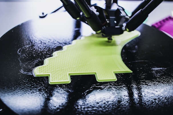 A 3D printer is in the process of creating a lime green object with a textured surface. The object being printed has a distinctive shape, possibly resembling a simplified dinosaur or creative design. The printer appears to be working on a circular black platform, reflecting light due to its glossy surface.