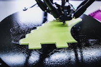A 3D printer is in the process of creating a lime green object with a textured surface. The object being printed has a distinctive shape, possibly resembling a simplified dinosaur or creative design. The printer appears to be working on a circular black platform, reflecting light due to its glossy surface.