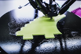 A 3D printer is in the process of creating a lime green object with a textured surface. The object being printed has a distinctive shape, possibly resembling a simplified dinosaur or creative design. The printer appears to be working on a circular black platform, reflecting light due to its glossy surface.