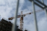 A construction crane towering over a building site with scaffolding, set against a partly cloudy sky. The crane is yellow and bears the name 'SDK'.