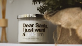 A scented candle with a humorous message sits on a shelf. The text on the candle reads 'Dear Santa, I just want a unicorn.' The area around the candle includes decorative elements such as a pine branch and a burlap wrap.