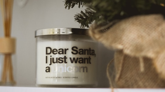 A scented candle with a humorous message sits on a shelf. The text on the candle reads 'Dear Santa, I just want a unicorn.' The area around the candle includes decorative elements such as a pine branch and a burlap wrap.