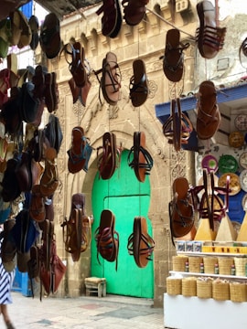 A colorful market scene features numerous hanging leather sandals in the foreground with a bright turquoise door on an ornately carved stone wall in the background. To the right, a market stall displays baskets and conical piles of spices. The environment appears vibrant and bustling, indicative of a traditional marketplace.