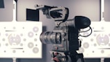 A professional video camera is mounted on a tripod, positioned in front of a technical test chart with various circles and patterns, typically used for calibrating cameras.