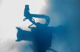 A silhouette of a video camera with a handle is projected against a backdrop of mist or fog. The image has a soft, bluish tint that gives it an ethereal and mysterious quality.