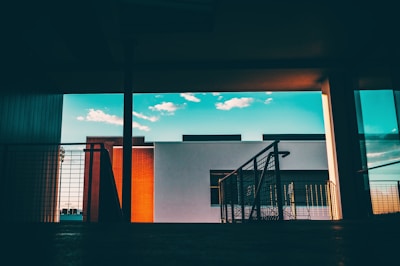 A modern architectural scene featuring a building with a minimalist design. The background includes a clear blue sky with scattered white clouds, while the foreground consists of a darkened interior space with railings and a hint of a shadowed floor.