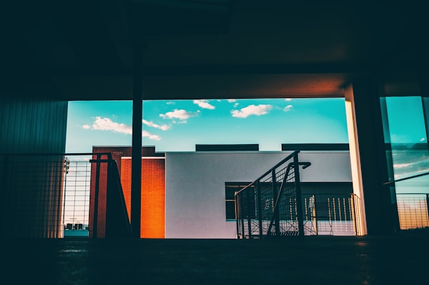 A modern architectural scene featuring a building with a minimalist design. The background includes a clear blue sky with scattered white clouds, while the foreground consists of a darkened interior space with railings and a hint of a shadowed floor.