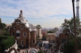 The image features two whimsical buildings with unique, organic shapes and textured surfaces, reminiscent of gingerbread houses. The architecture is decorated with white ceramic details and has a fairytale-like appearance. The scene is set in a park with trees, while groups of people walk and gather in the area.