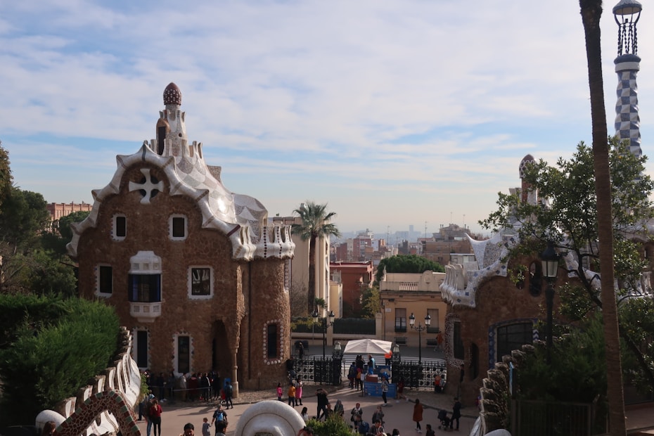 The image features two whimsical buildings with unique, organic shapes and textured surfaces, reminiscent of gingerbread houses. The architecture is decorated with white ceramic details and has a fairytale-like appearance. The scene is set in a park with trees, while groups of people walk and gather in the area.