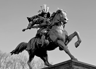A dramatic bronze statue depicts a samurai warrior in full armor riding a rearing horse. The warrior is brandishing a sword, and both figures are detailed and dynamic against a clear sky.