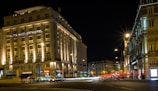A nighttime cityscape featuring a well-lit hotel on a busy street corner, with light trails from passing cars. The architecture is classic and ornate, suggesting a historic building. Street lamps illuminate the area, casting a warm glow on the surroundings.