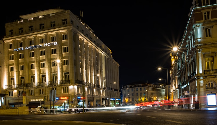A nighttime cityscape featuring a well-lit hotel on a busy street corner, with light trails from passing cars. The architecture is classic and ornate, suggesting a historic building. Street lamps illuminate the area, casting a warm glow on the surroundings.