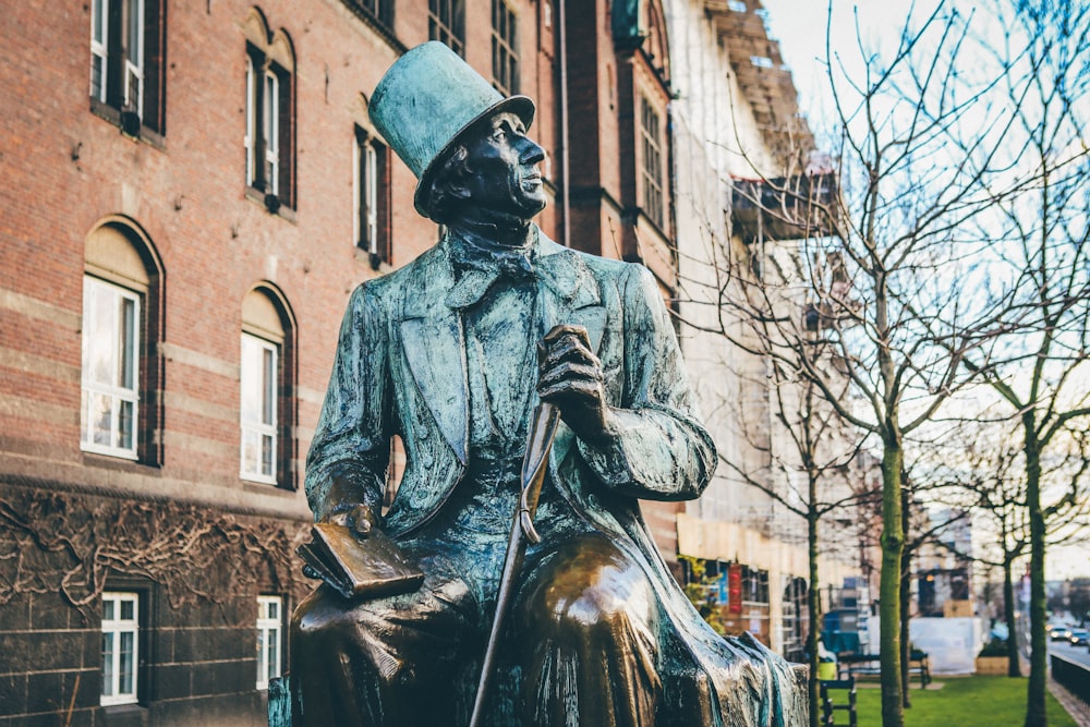 모자와 코트를 입은 남자가 책과 지팡이를 들고 동상에 앉아 있습니다.